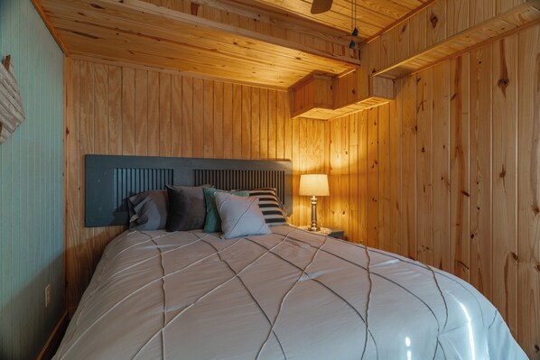 Une chambre à coucher aux murs lambrissés, avec un lit, des oreillers et une table de chevet surmontée d’une lampe.
