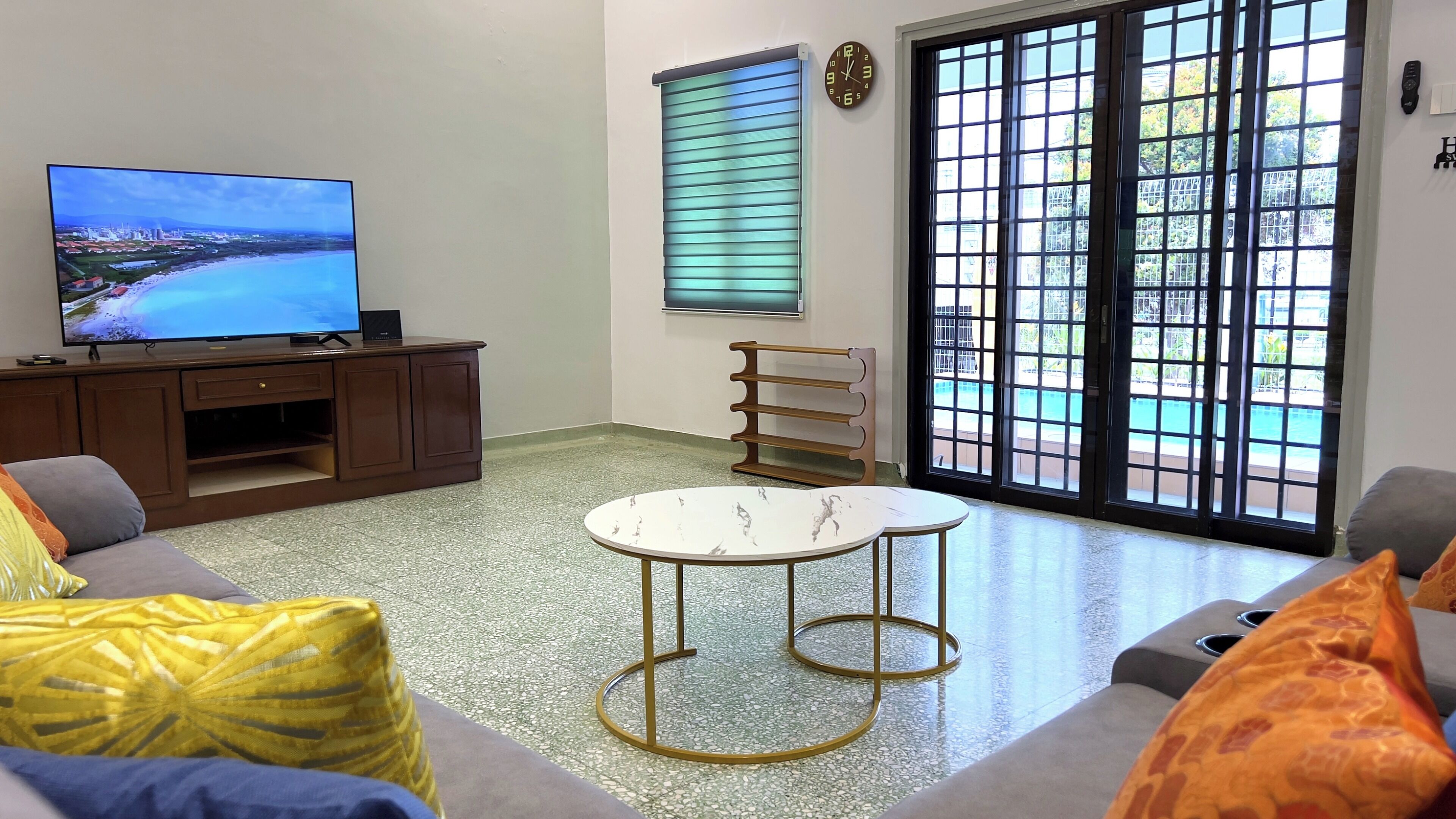 Uma sala de estar com televisão, sofá, mesa de centro e vista para uma piscina através de grandes janelas.