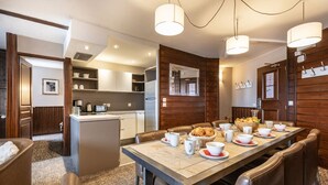 Ein moderner Essbereich mit einem langen Tisch, gedeckt für sechs Personen, umgeben von Ledersesseln. Im Hintergrund ist eine Küche mit einer integrierten Kochinsel und einem Kühlschrank erkennbar.