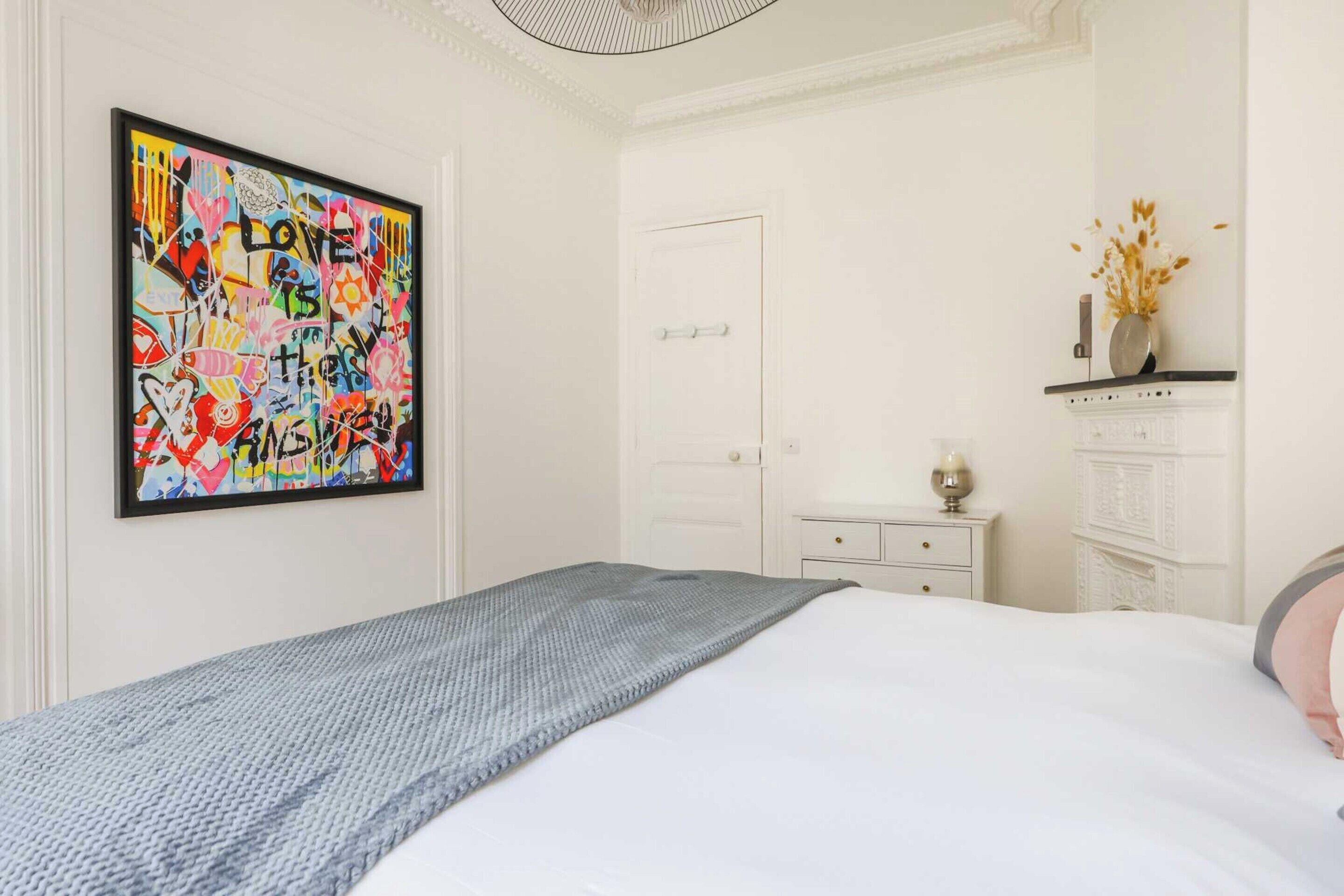 A bedroom with a bed, a white door, a small bedside table with a lamp, and a decorative vase on a shelf.