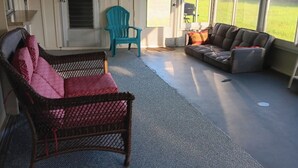 Un porche techado con un ventilador de techo, una silla de mimbre con cojines rojos, una silla Adirondack turquesa y un sofá seccional.
