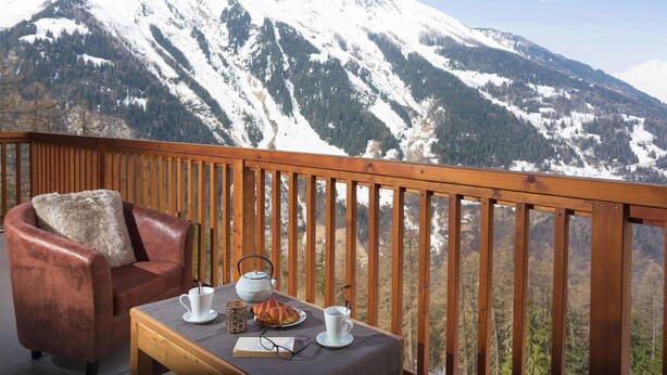 A balcony with a leather chair, a table with a teapot, cups, and a croissant, overlooking snow-covered mountains. - Apartment in Sainte-foy With Ski-in/ski-out (Sainte-Foy-Tarentaise)
