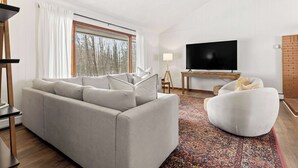 A modern living room with a sofa, a white armchair, a flat-screen TV, and a patterned rug. - Scenic Escape Kayaks Game Room on M22 (Frankfort)