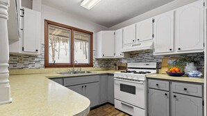 A kitchen with white cabinets, a built-in stove, and a window with a view of trees. - Scenic Escape Kayaks Game Room on M22 (Frankfort)