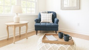 A room with a blue armchair, a small wooden side table with a white vase, a tray with a book and two black mugs, and a large window with a view of trees. - Charming & Spacious 4 BR w/ Private Backyard in lovely Summerville (Summerville)