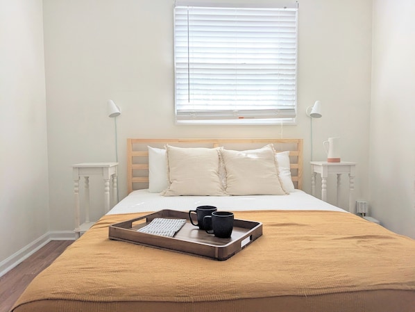 A neatly made bed with a wooden headboard, a beige throw blanket, and a tray with a black mug and a white cup. There are two bedside lamps on white nightstands, and a window with blinds above the bed.