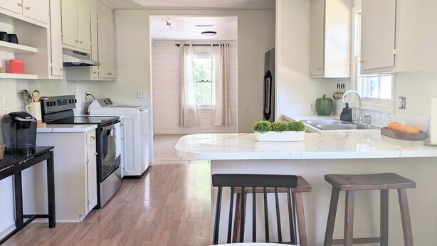 A modern kitchen with white cabinetry, a central island, and a dining area with a white table and wooden chairs. - Charming & Spacious 4 BR w/ Private Backyard in lovely Summerville (Summerville)