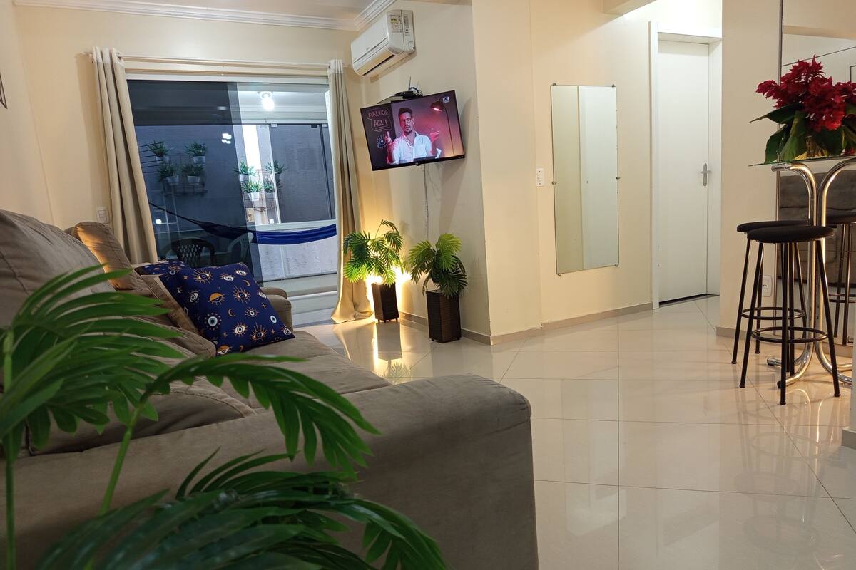 A living room with a sofa, a TV mounted on the wall, a dining area with a bar stool, and potted plants.