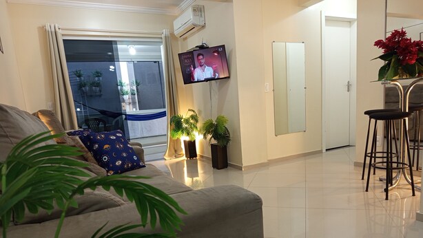 A living room with a sofa, a TV mounted on the wall, a dining area with a bar stool, and potted plants. - Apto 102 Ed Gabriel na divisa do Parque (Piratuba)