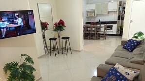 A modern living room with a TV, a dining area with stools, and a kitchen in the background. - Apto 102 Ed Gabriel na divisa do Parque (Piratuba)