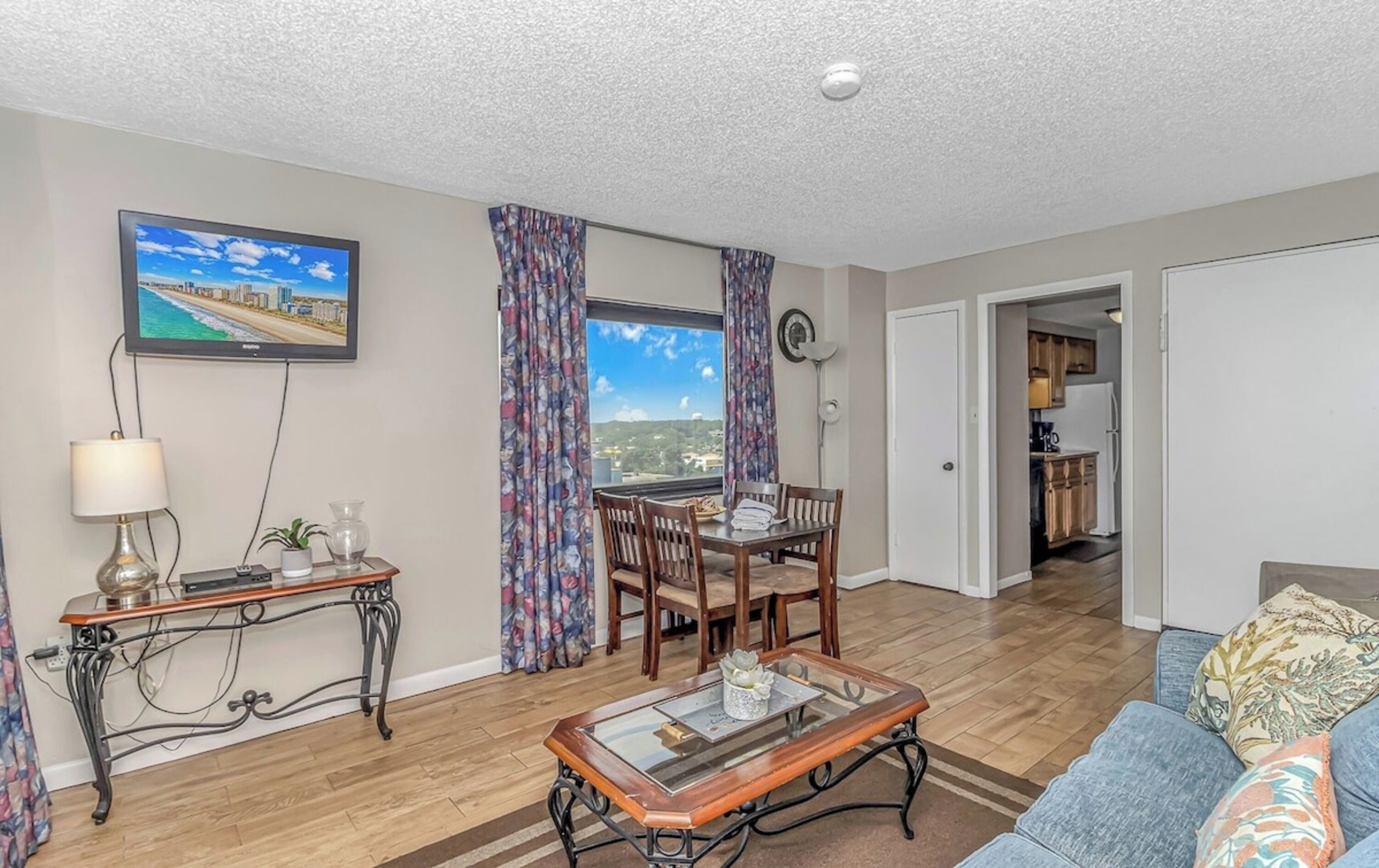 A living room with a dining table, a sofa, a TV mounted on the wall, and a view of the city through the window.