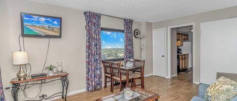 A living room with a dining table, a sofa, a TV mounted on the wall, and a view of the city through the window.