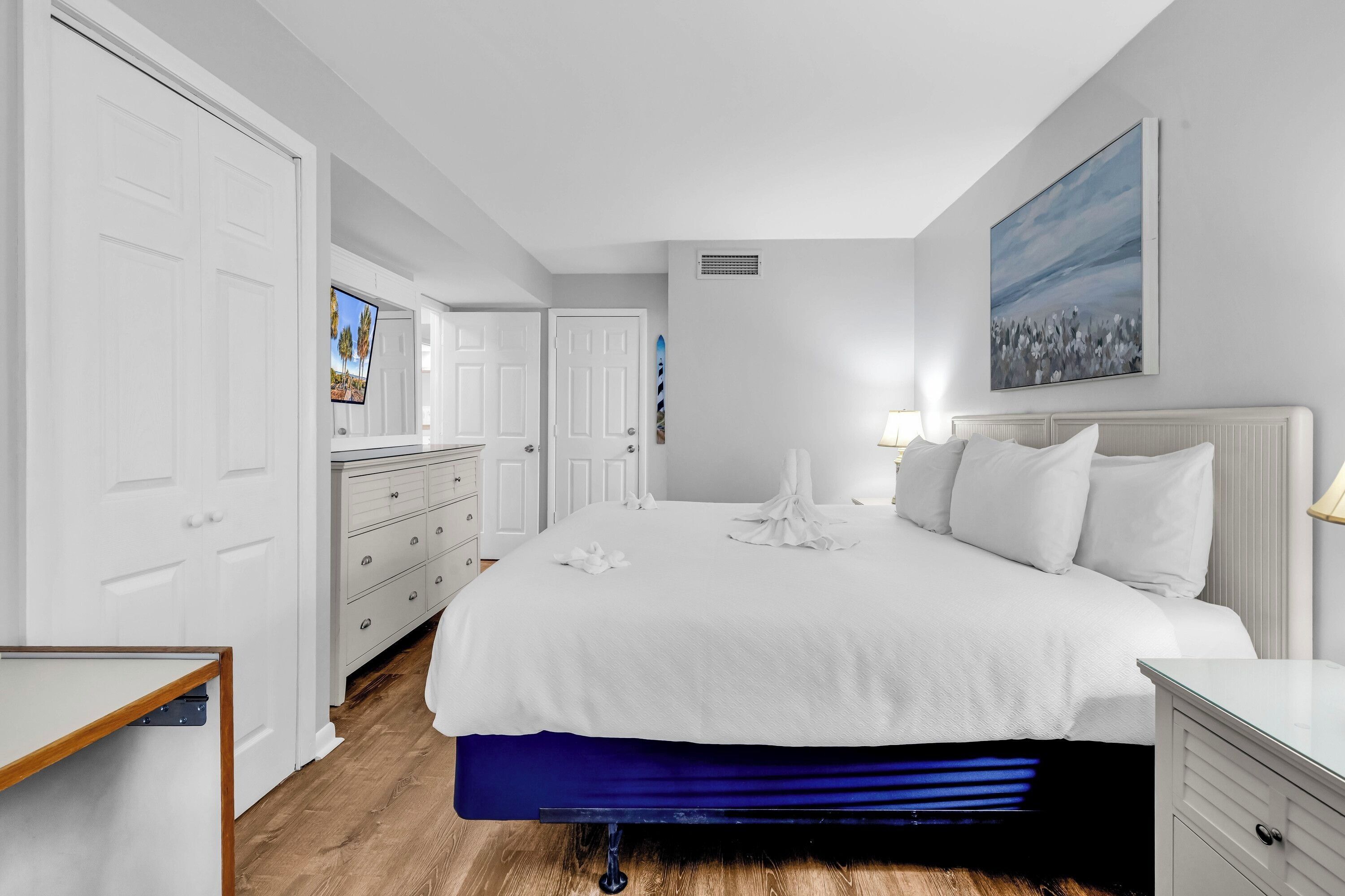 A bedroom with a large bed, white walls, a painting, and a TV on a dresser.