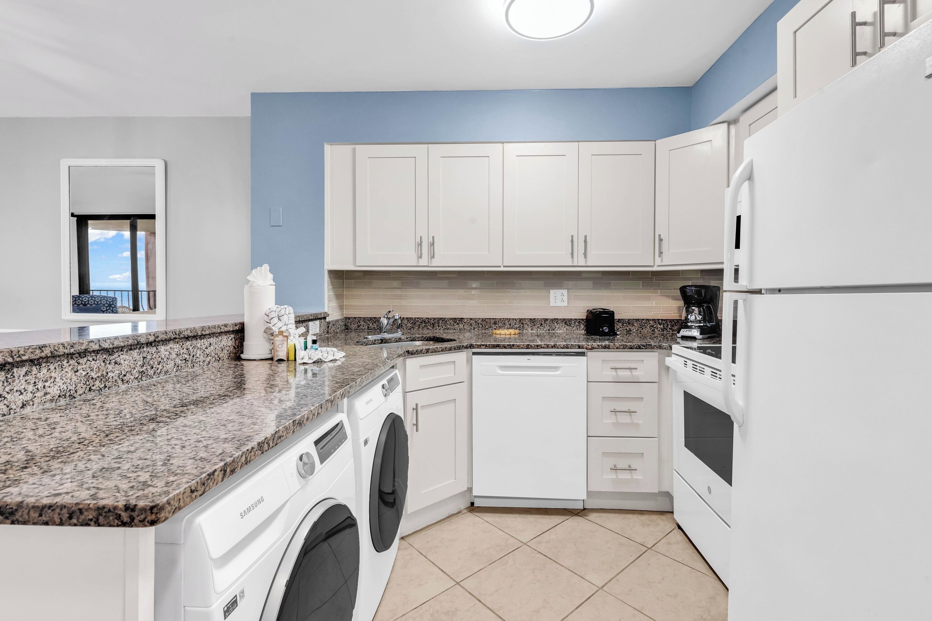 A modern kitchen with white cabinets, a granite countertop, a washing machine, a dishwasher, and a refrigerator.