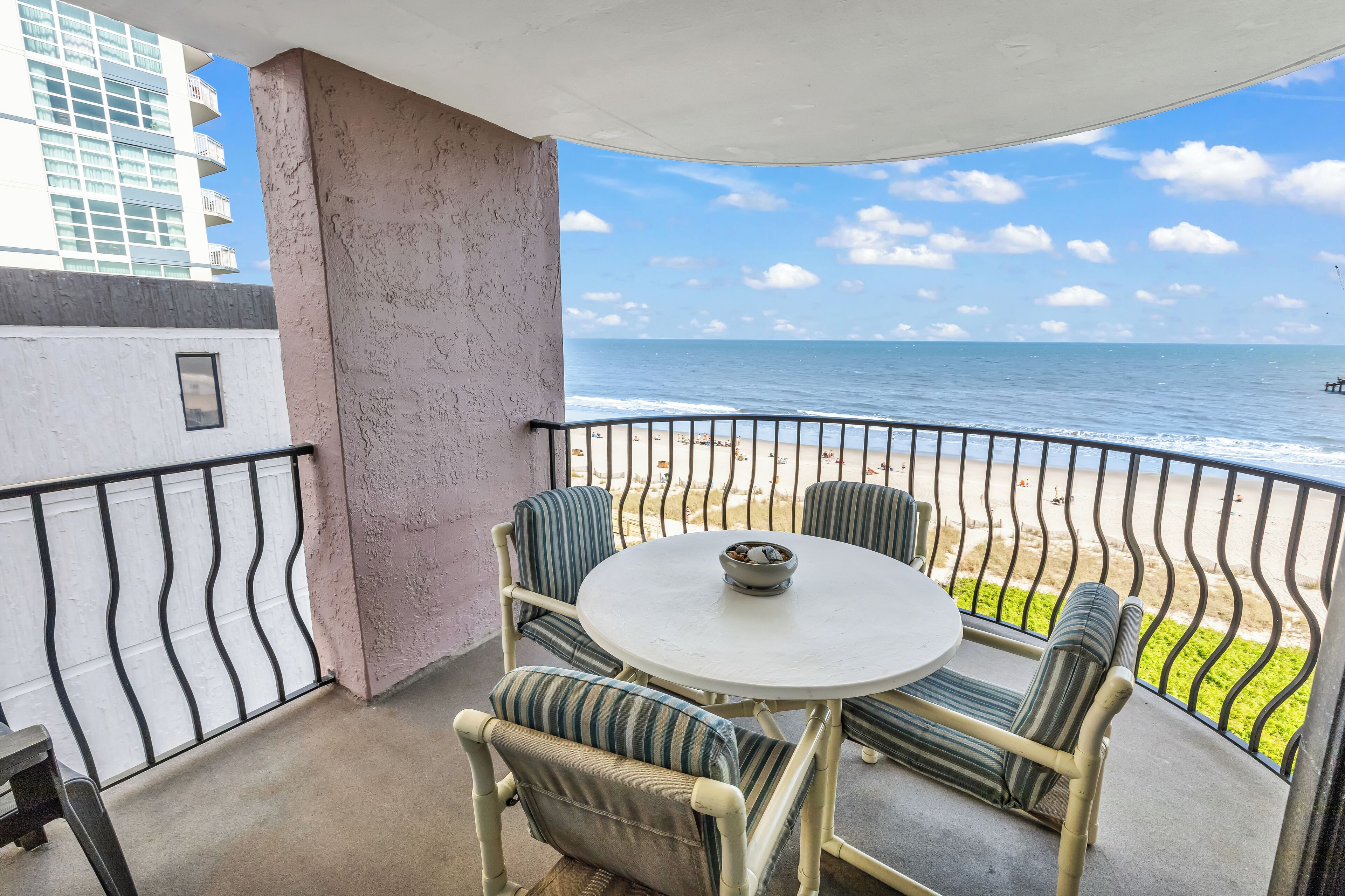 A balcony with a round table and chairs, overlooking a beach with a clear sky and ocean.