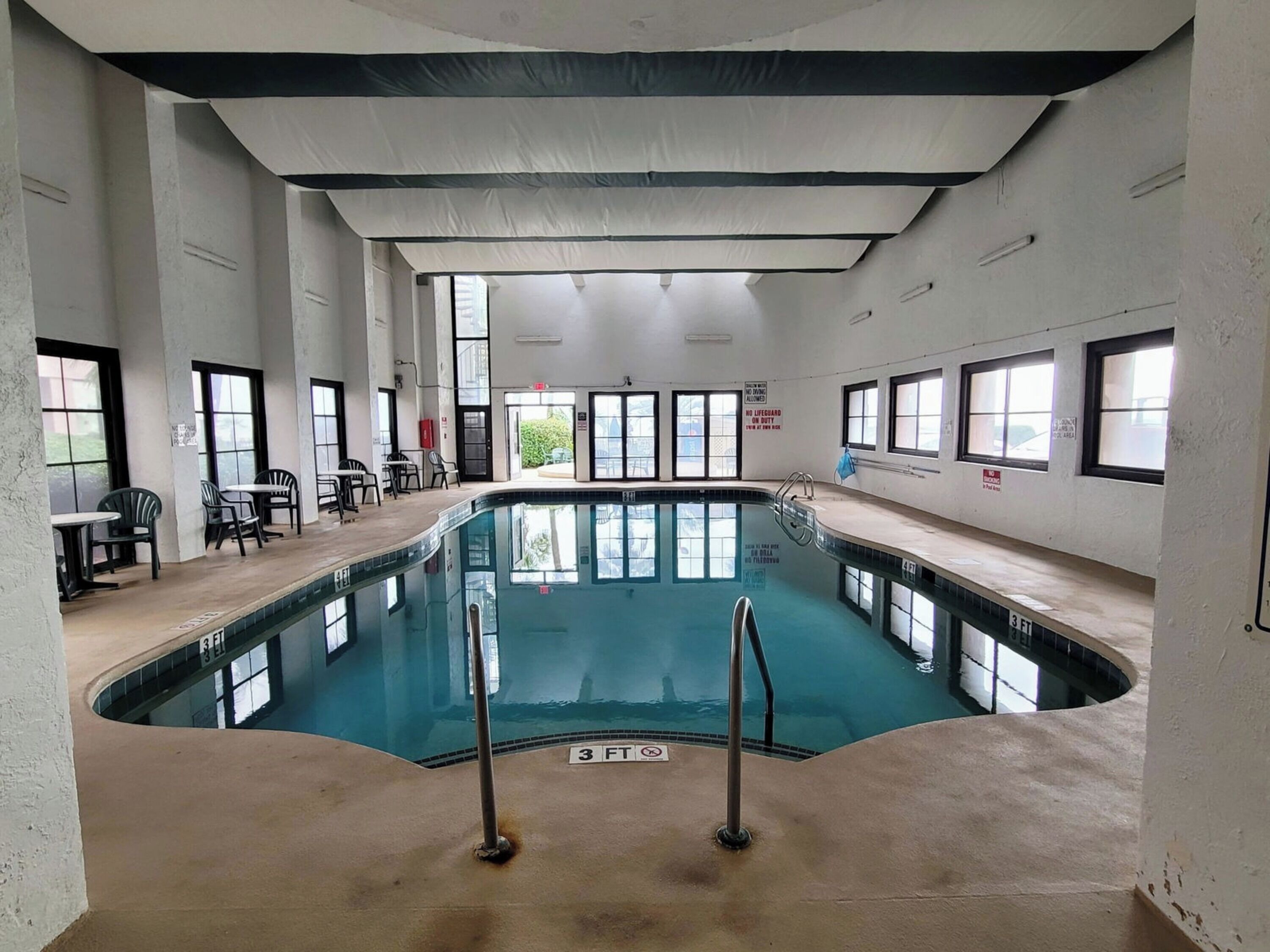 An indoor swimming pool with clear water, surrounded by walls with windows. There are chairs and tables outside the pool area.