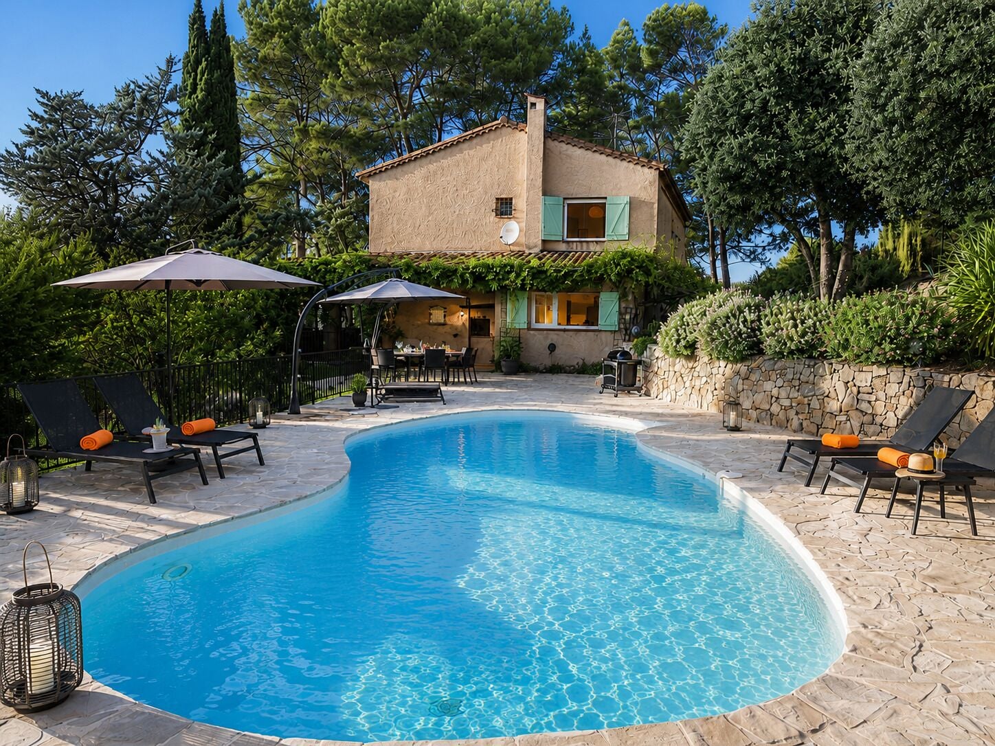 A swimming pool with lounge chairs and umbrellas, surrounded by a stone patio and a building with a stone wall and greenery.