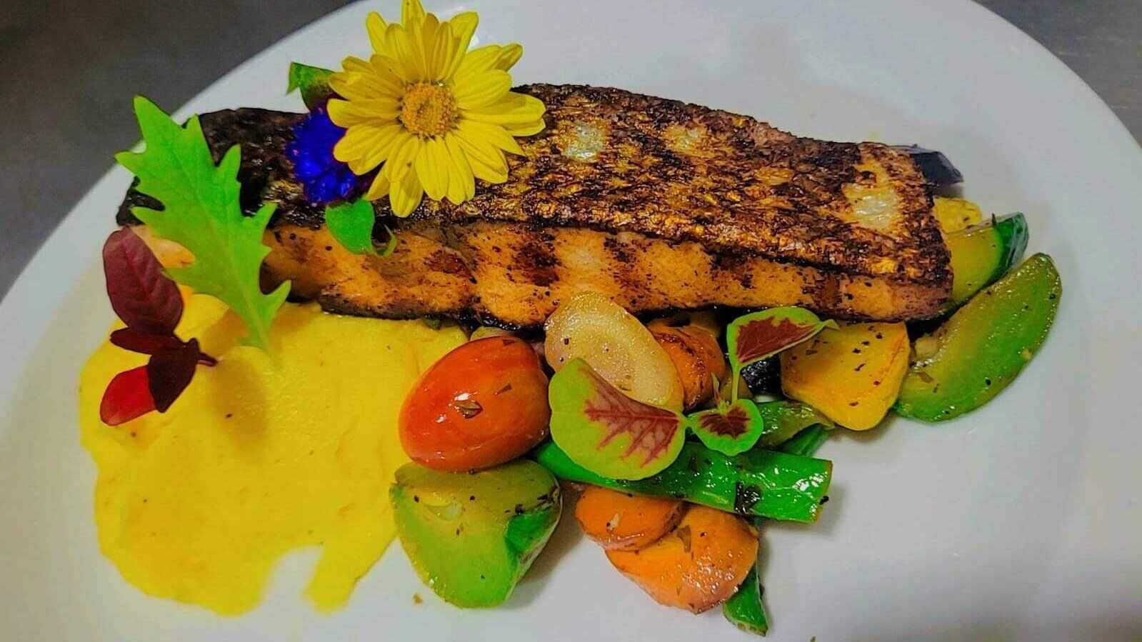 Een bord met gegrilde vis, geroosterde groenten en een gele saus.