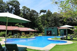 A swimming pool with lounge chairs and umbrellas, surrounded by greenery and buildings with red roofs. - Hacienda Barú Lodge (Savegre)