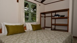 A bedroom with a bed, two pillows, a window with curtains, and a wooden shelf unit. - Hacienda Barú Lodge (Savegre)