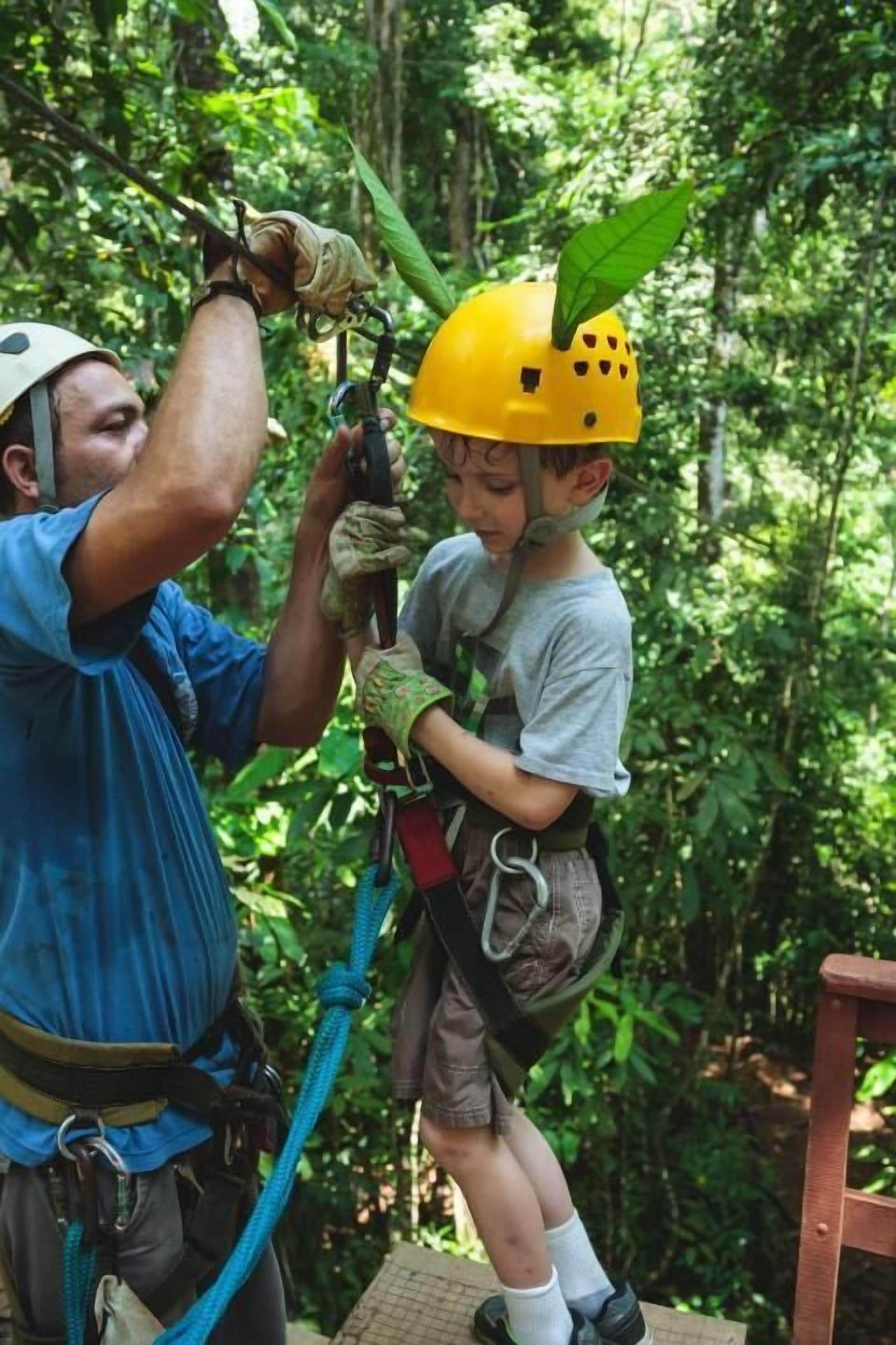 A person assisting another with a safety harness in a forested area.