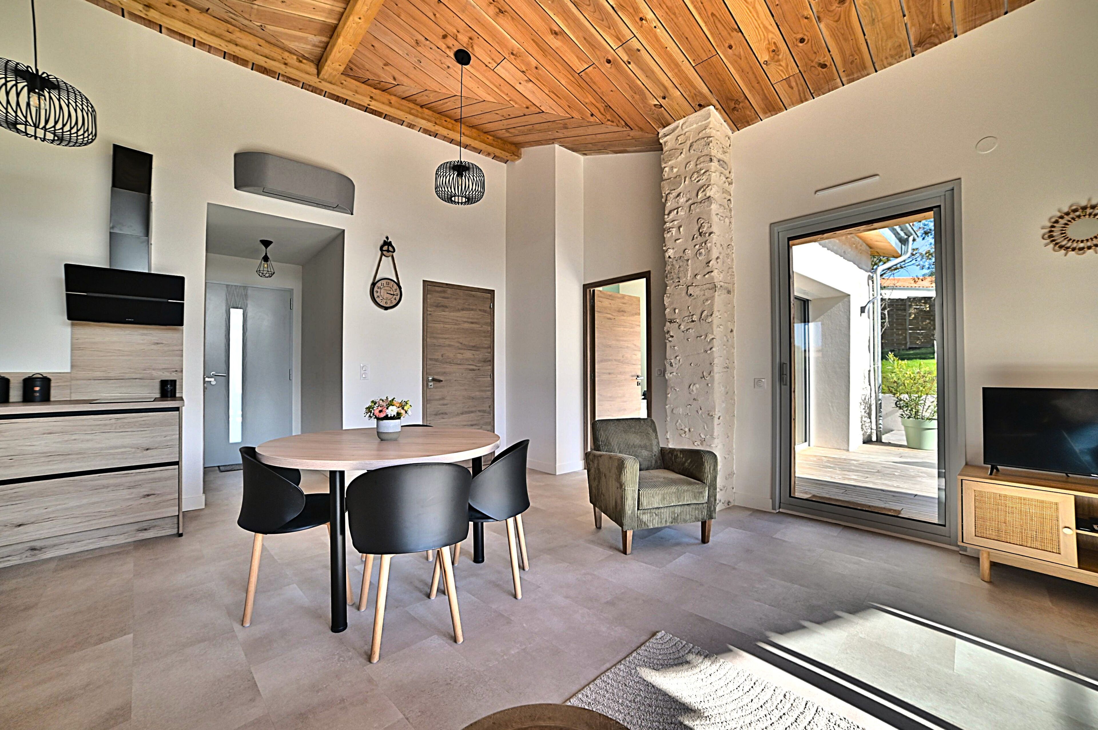 A modern kitchen with a dining area, wooden ceiling, stone pillar, and a TV in a wooden cabinet.