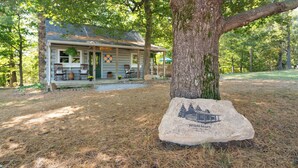 A rustic cabin with a porch, surrounded by trees and a stone with a house and trees engraving.