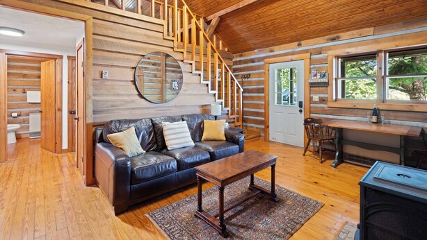 A log cabin interior with a wooden staircase, leather sofa, dining table, and a large window.