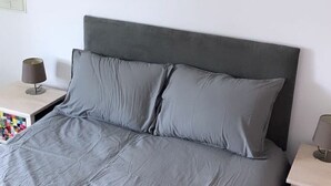 Un lit avec une tête de lit grise et des oreillers, une table de chevet avec une lampe, et un interrupteur fixé au mur.