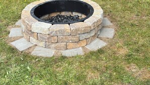 A circular stone fire pit surrounded by a stone border, set in a grassy area with a wooden bridge in the background. - TRANQUIL RETREAT IN GRAYLAND - Minutes from Westport's Coast (Grayland)