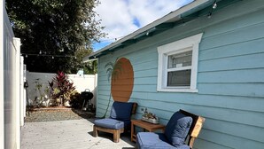 A covered patio with two wooden chairs and a small table, a decorative wall with a palm tree, and a white fence in the background. - Charming bungalow studio between the beach and downtown in welcoming St. Pete (St. Petersburg)