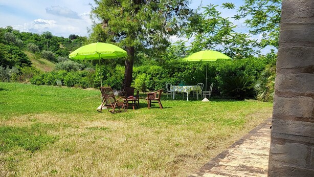 A patio area with outdoor furniture and umbrellas, a grassy field, and a stone pillar.