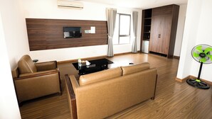 A modern living room with a sofa, a coffee table, a TV mounted on a wooden wall, a window with curtains, and a fan. - Kim Thanh Hotel (Vinh Phuc)