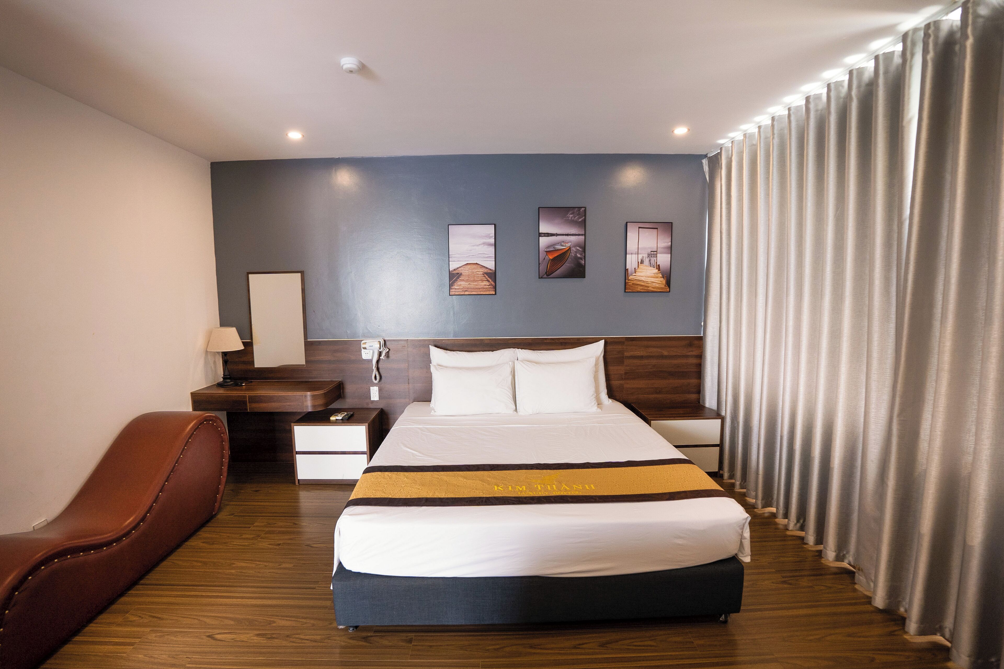 A hotel room with a large bed, a wooden nightstand, a desk, a brown leather chair, and a view of the sea through the curtains.