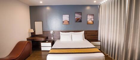 A hotel room with a large bed, a wooden nightstand, a desk, a brown leather chair, and a view of the sea through the curtains.