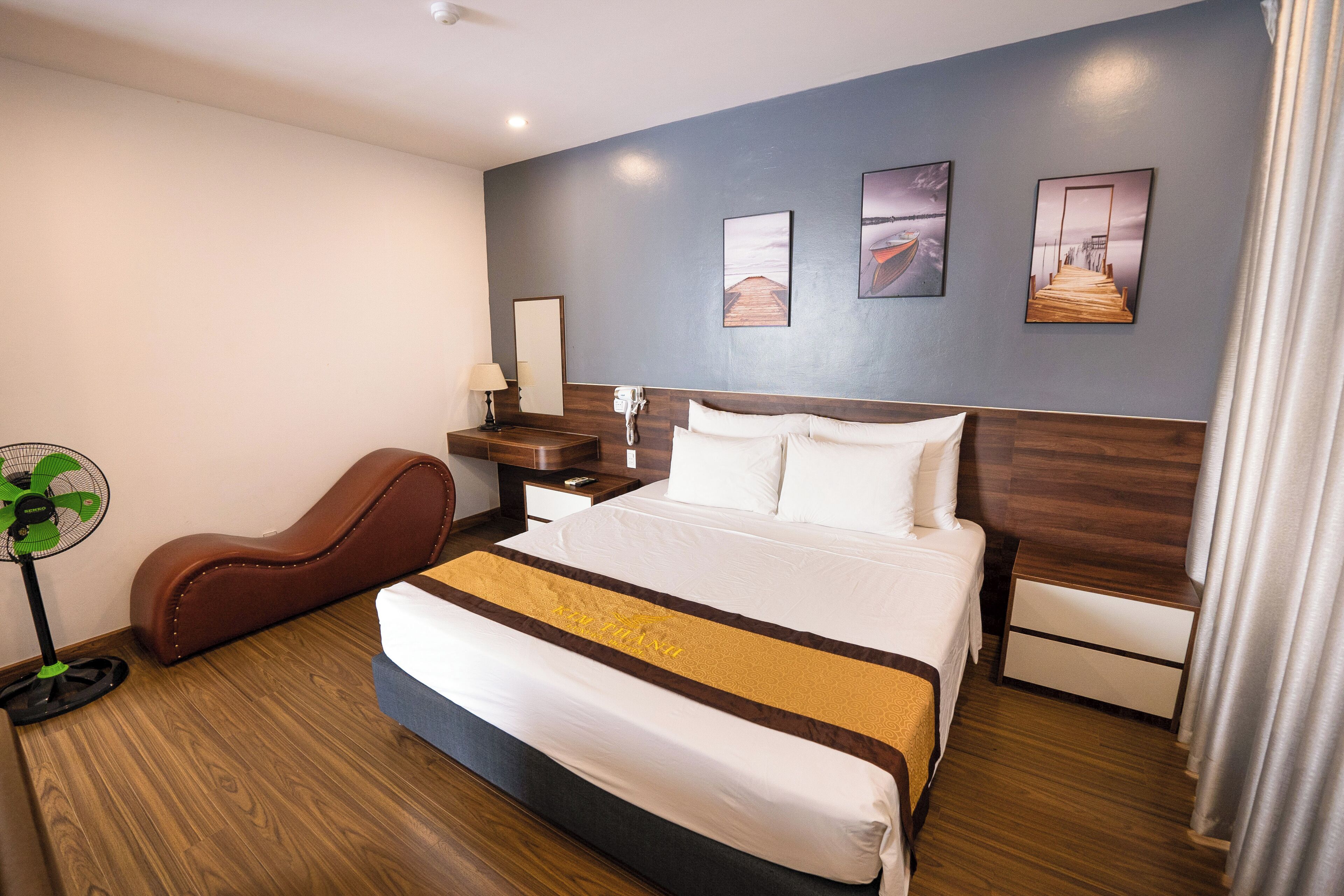 A hotel room with a large bed, a wooden headboard, a desk with a lamp, a chair, and a wall-mounted fan.