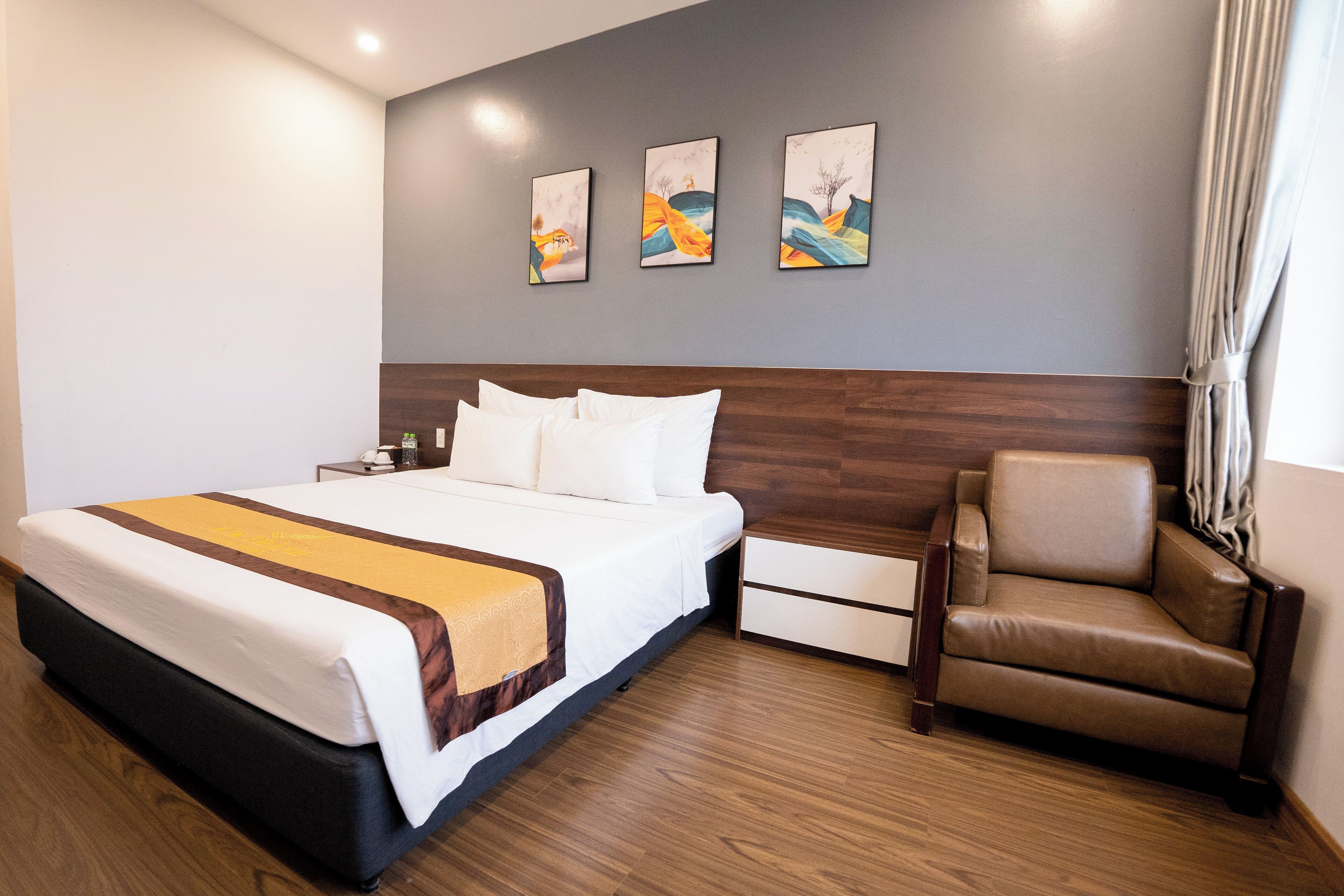 Quarto de hotel com uma cama grande, cabeceira de madeira, uma poltrona de couro marrom, mesa de cabeceira e três quadros na parede.