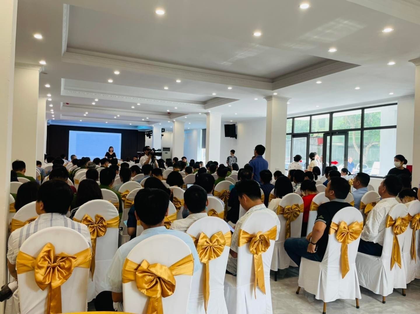 Un salón de conferencias con asistentes sentados frente a un escenario, sillas blancas con cintas doradas y amplias ventanas.