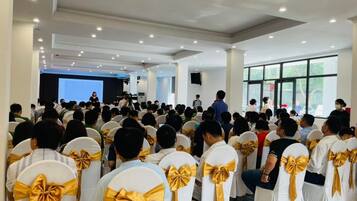 Un salón de conferencias con asistentes sentados frente a un escenario, sillas blancas con cintas doradas y amplias ventanas.