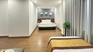 A modern hotel room with a wooden floor, a bed with a white and gold bedspread, a bedside table with a lamp, and a wall-mounted fan. - Kim Thanh Hotel (Vinh Phuc)