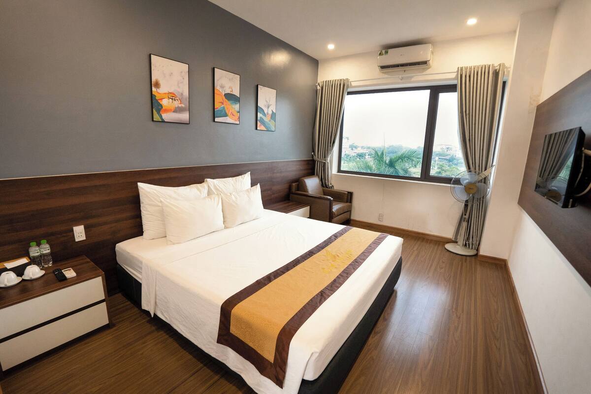 Habitación de hotel con una cama grande, una mesita de noche con lámpara, una silla, un ventanal con cortinas y una obra de arte en la pared.