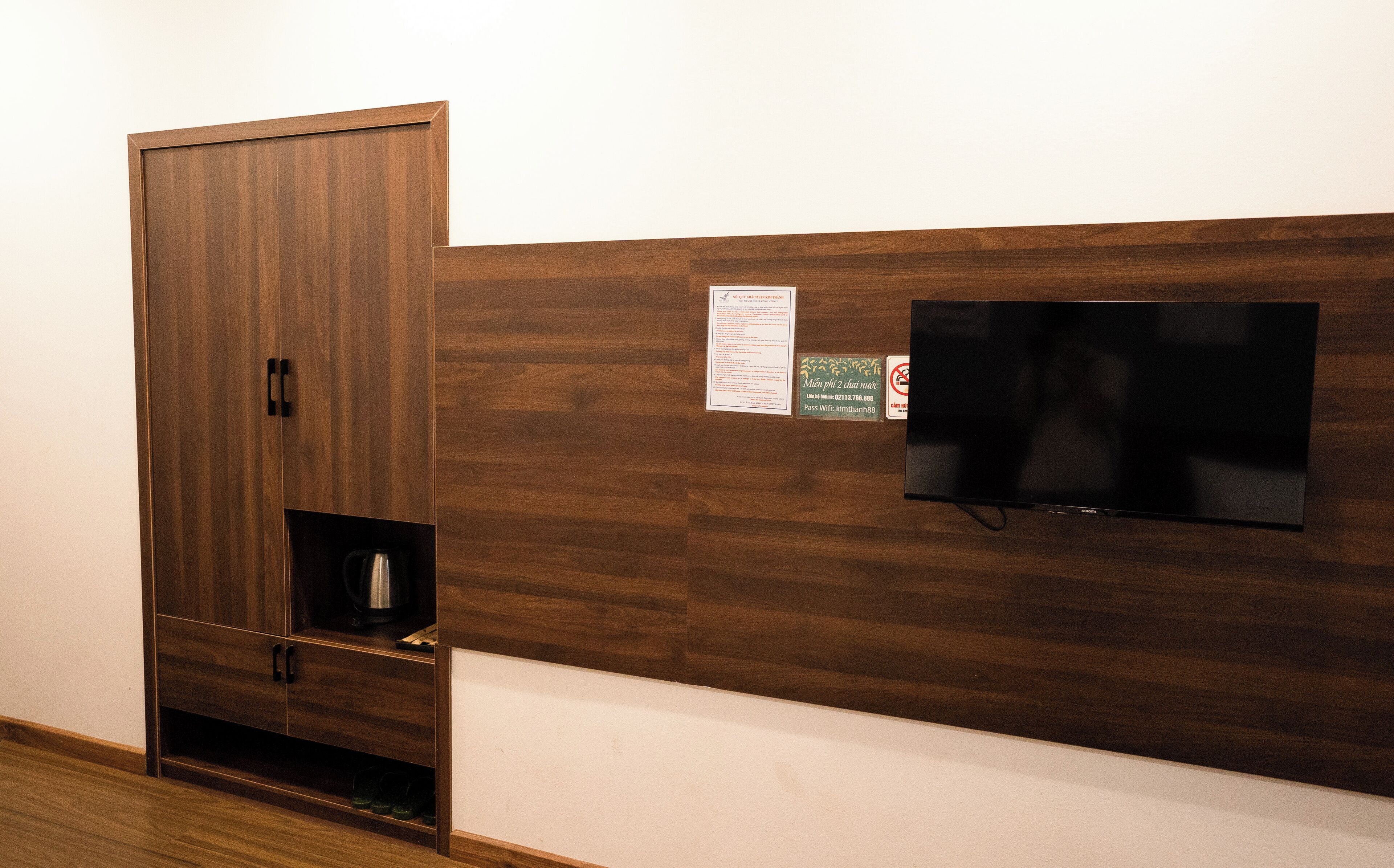 A room with a wooden cabinet, a television mounted on the wall, and a small shelf with a kettle.