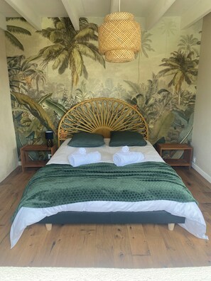 Un letto con copriletto verde, due cuscini e una testiera in rattan. Sono presenti due comodini con lampade e un affresco floreale tropicale sulla parete.