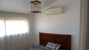 A bedroom with a wooden headboard, a bed with a colorful quilt, a checkered pillow, a hanging lantern, and an air conditioning unit on the wall.