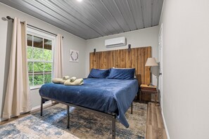 Une chambre à coucher comprenant un lit, une fenêtre, un appareil de climatisation, une lampe et une tête de lit en bois.
