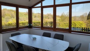 Un comedor con una mesa blanca y seis sillas, ventanas amplias con vistas a un paisaje verde y un tragaluz arriba.