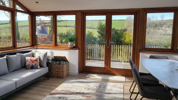 Una habitación iluminada por el sol con un sofá, una mesa de comedor y vistas a un campo verde a través de amplias ventanas.