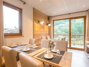 Comedor con mesas dispuestas para cuatro, paredes de madera y grandes ventanas que ofrecen vistas a un paisaje verde.