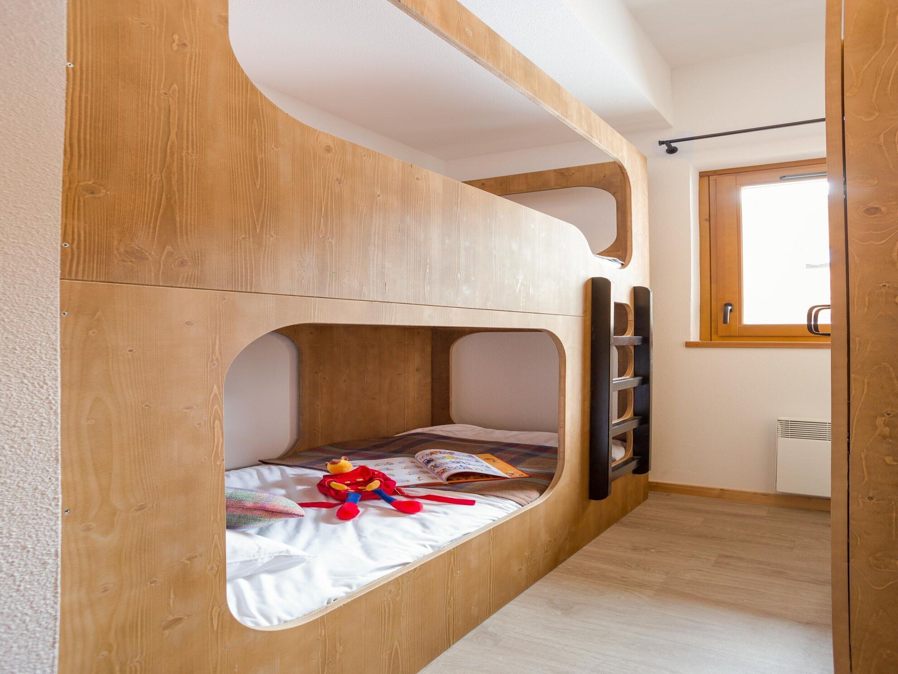 A bunk bed with a wooden finish, a window with a wooden frame, and a radiator on the wall.