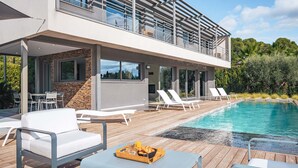 Un área de piscina moderna al aire libre con sillones de descanso y una mesa, rodeada por una terraza de madera y un edificio de dos pisos con grandes ventanales.